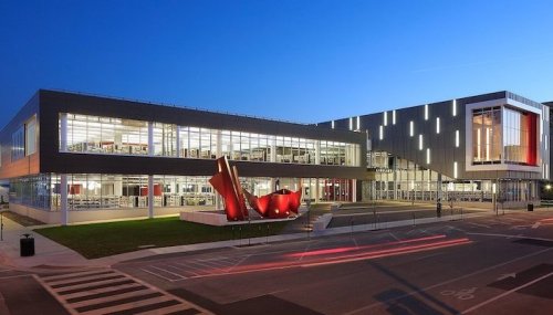 Modern building with large glass windows, red accents, and a striking red sculpture in front, illuminated at dusk.