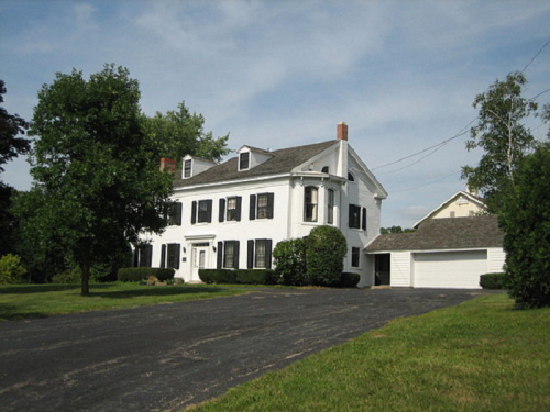 A white two-story house with black shutters, surrounded by greenery and a paved driveway leading to a garage.