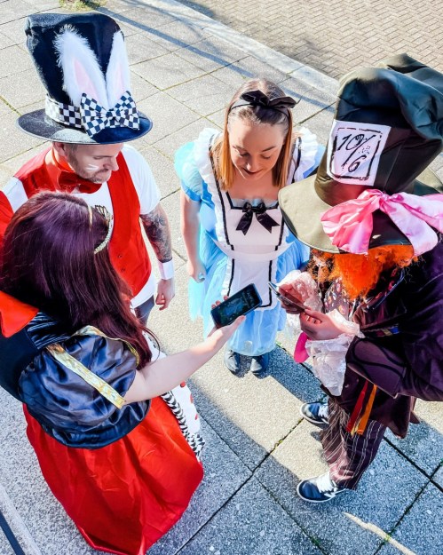 A group of four people in colorful costumes, including Alice and the Mad Hatter, looking at a phone together.