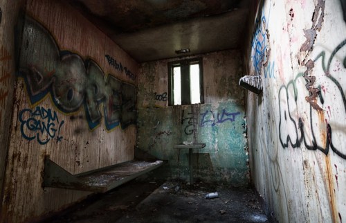 An abandoned room with peeling walls, graffiti, and a rusted bed frame, illuminated by a small window.