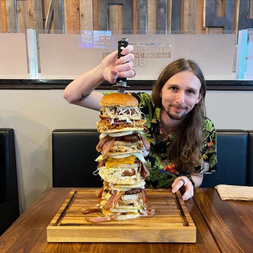 A person with long hair holds a towering stack of burgers topped with various ingredients in a restaurant setting.