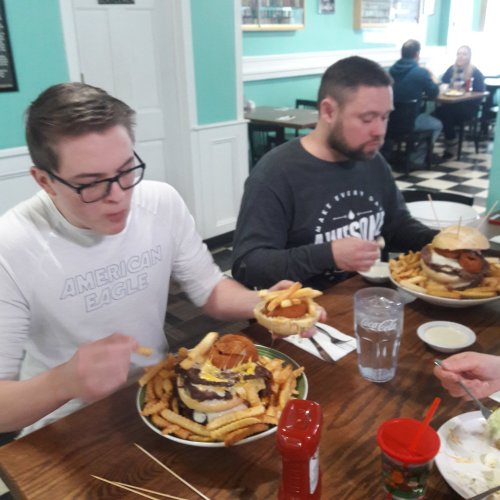 Two men enjoy large burgers and fries at a restaurant, with one man taking a bite and the other looking at his meal.