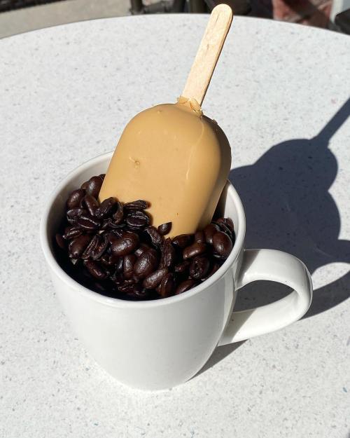 A white mug filled with coffee beans, topped with a coffee-flavored popsicle on a sunny surface.