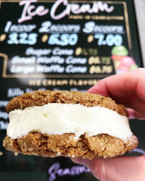 A hand holding a cookie ice cream sandwich with a menu board in the background featuring ice cream prices.