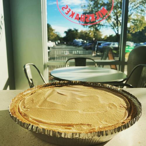 A creamy pie in a silver tin sits on a table, with a bright window view of a parking lot and trees in the background.
