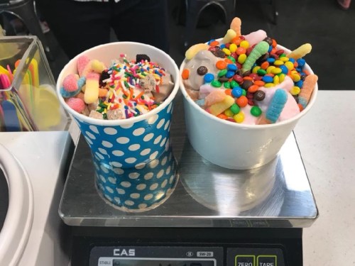 Two colorful ice cream cups topped with sprinkles, candy, and gummy treats, placed on a scale.