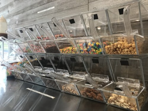 A display of clear containers filled with various toppings like sprinkles, nuts, and candies on a wooden background.
