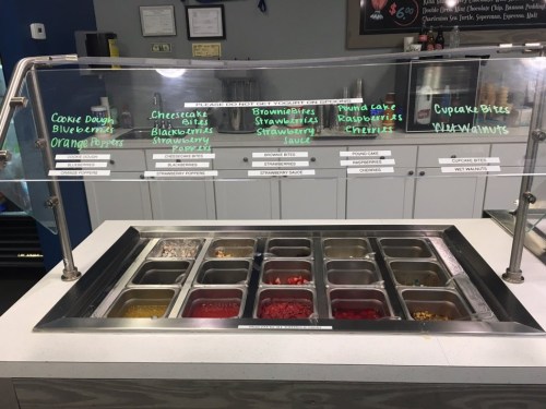 A dessert toppings bar with various options like cookie dough, fruits, and sauces in labeled containers.