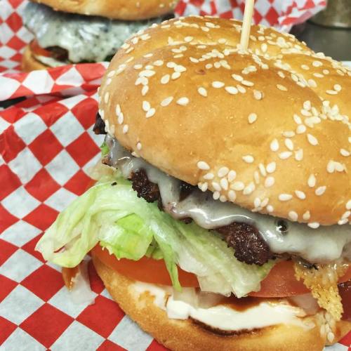 A close-up of a sesame seed burger with lettuce, tomato, and melted cheese, served on a red and white checkered paper.