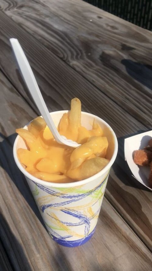 A cup of fries topped with melted cheese, served with a plastic spoon on a wooden table.