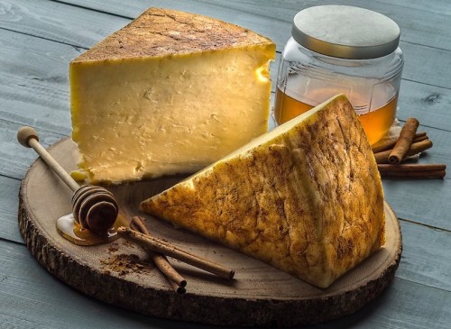 A wedge of cheese on a wooden board, accompanied by honey, cinnamon sticks, and a jar of honey.