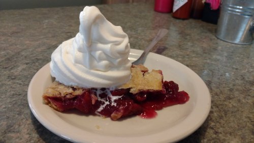 A slice of cherry pie topped with a generous swirl of whipped cream on a white plate.