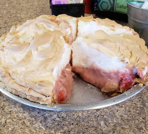 A pie with a golden meringue topping, partially sliced to reveal a pink filling and a flaky crust on a silver pie plate.