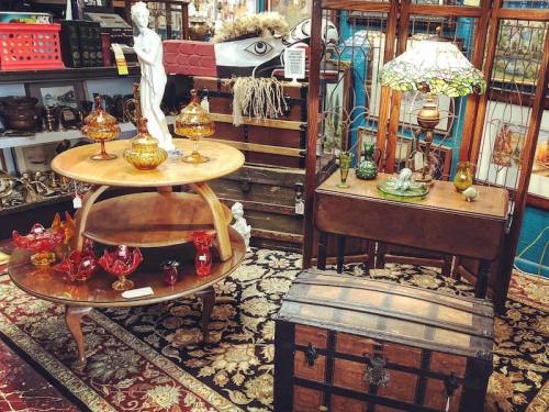 A vintage shop display featuring wooden tables, glassware, a statue, and a decorative lamp on a patterned rug.