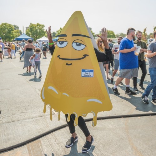 A person in a large cheese wedge costume waves at a busy outdoor event with people in the background.