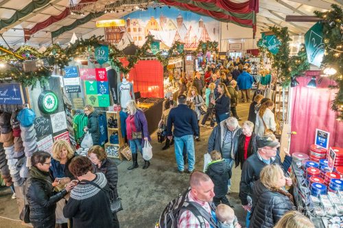 A bustling indoor market filled with people browsing stalls decorated for the holidays, with festive lights and banners.
