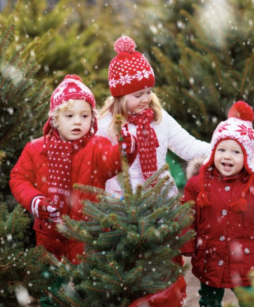 Three children in festive winter attire play among Christmas trees, surrounded by falling snow.