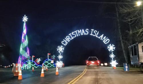 A festive archway reading "Christmas Island" with colorful lights, a decorated tree, and traffic cones at night.
