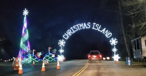 A festive archway reading "Christmas Island" with colorful lights and a decorated tree beside a car on a dark road.