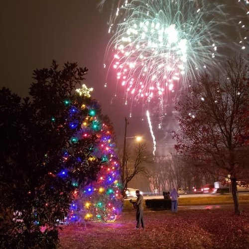 A brightly lit Christmas tree with colorful lights, fireworks bursting in the night sky above.