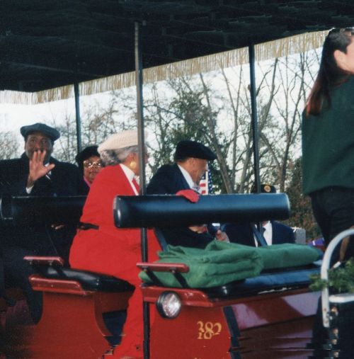 A festive parade scene with people in winter attire, some waving, seated on a decorated float under a canopy.
