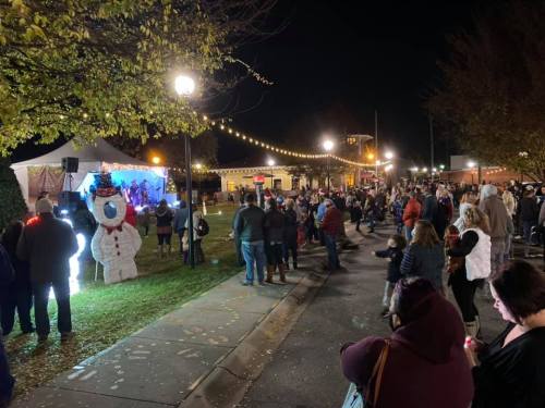 A crowd gathers at night for a festive event, with a stage and a snowman decoration in a park.