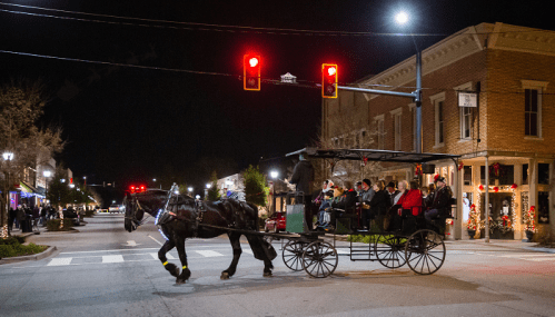 A horse-drawn carriage with passengers crosses a street at night, illuminated by traffic lights and festive decorations.
