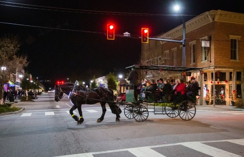 A horse-drawn carriage with passengers crosses a street at night, illuminated by traffic lights and festive decorations.