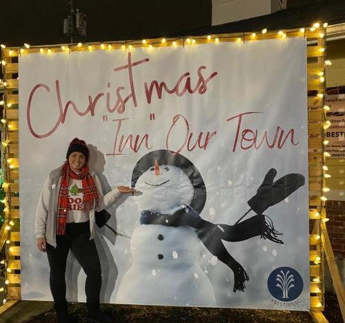 A person poses next to a festive backdrop featuring a snowman and the text "Christmas 'Inn' Our Town," decorated with lights.