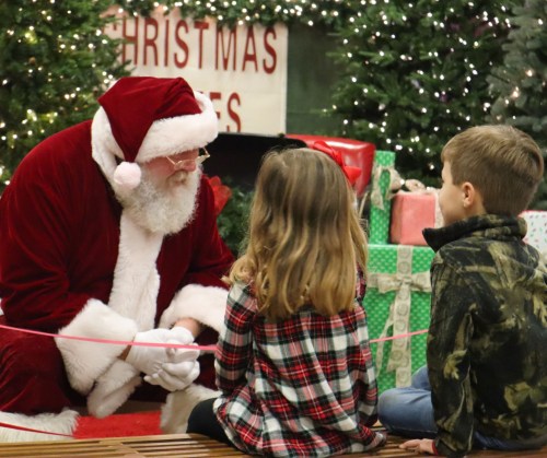 Santa Claus sits on a bench talking to two children in front of a festive backdrop with Christmas decorations.
