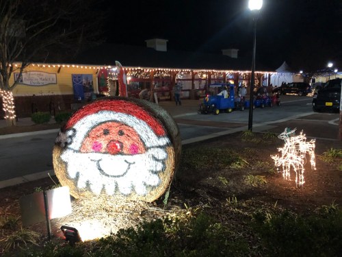 A festive scene featuring a hay bale painted like Santa and a light-up reindeer, with holiday lights in the background.