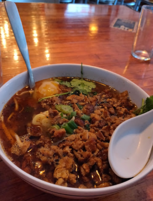 A bowl of soup with noodles, minced meat, herbs, and a soft-boiled egg, served with a spoon on a wooden table.