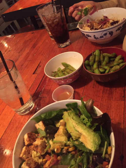 A table with a salad bowl, edamame, and drinks, with a hand reaching for a bowl of food in the background.