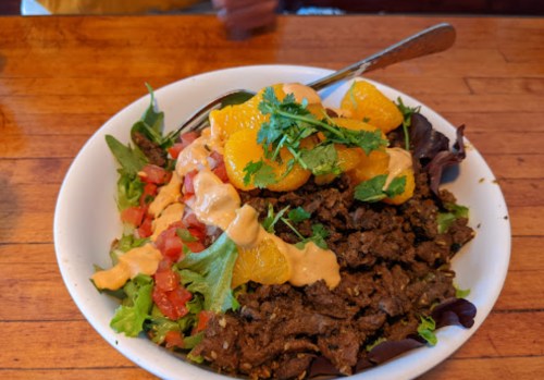 A bowl of salad topped with seasoned meat, orange segments, tomatoes, and a creamy dressing, garnished with cilantro.
