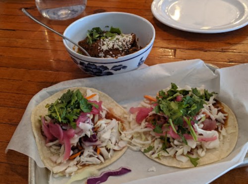 Two tacos topped with cabbage, pickled onions, and cilantro, alongside a bowl of mole with a sprinkle of cheese.