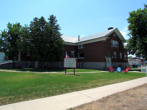 A brick building with a metal roof, surrounded by trees and a grassy area, featuring red chairs and a sign in front.