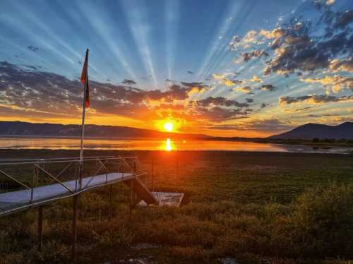 A vibrant sunset over a calm lake, with rays of light breaking through clouds and a small dock in the foreground.