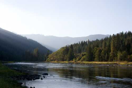 A serene river flows through a valley surrounded by mountains and lush green trees under a clear sky.