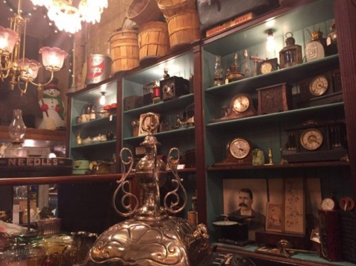 A vintage shop display featuring shelves of antique clocks, glassware, and decorative items under warm lighting.