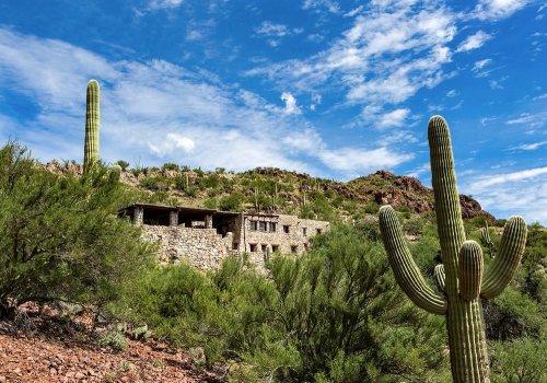 A stone building nestled in a desert landscape, surrounded by cacti and rocky hills under a blue sky with clouds.