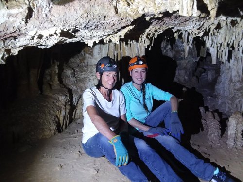 Two women in helmets sit on the cave floor, surrounded by rock formations and dim lighting.