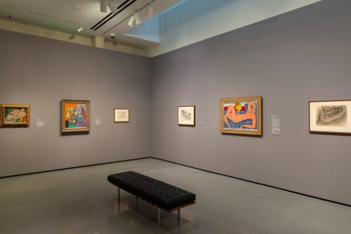 A gallery wall displaying various framed artworks, with a black bench in the center and soft lighting.