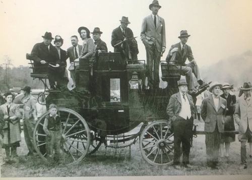 A vintage photo of a large group of people, some on a horse-drawn carriage, dressed in early 20th-century attire.