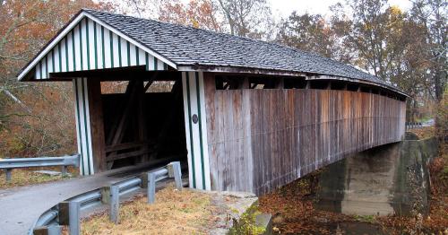 A rustic covered bridge with a wooden structure and green-striped roof, surrounded by autumn foliage.