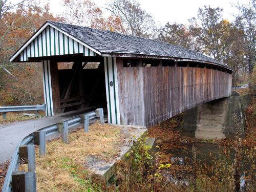 A wooden covered bridge with a sloped roof, surrounded by autumn foliage and a small stream.