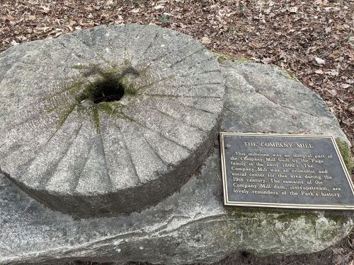 A stone millstone with a plaque about the Company Mill's history, surrounded by leaves and trees.