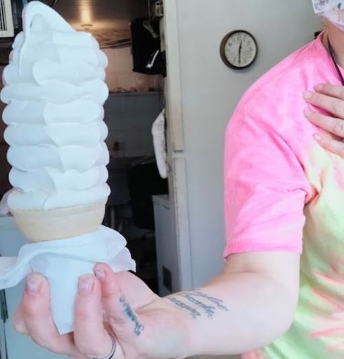 A person holds a tall, swirled soft serve ice cream cone with a colorful shirt and a mask in a casual setting.