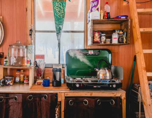 A cozy kitchen with a camping stove, kettle steaming, shelves of spices, and a wooden ladder in a rustic setting.