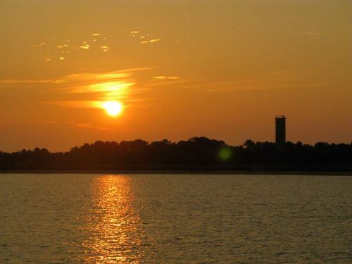 Sunset over a calm water body, with a silhouette of a tower and trees against a golden sky.