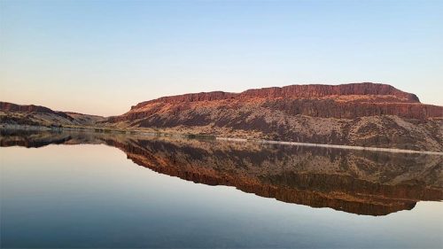 A serene landscape featuring a calm lake reflecting a rocky, layered cliff under a clear sky.